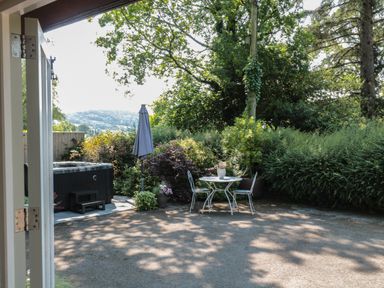An outdoor patio area with a small table and two chairs near shrubs and trees at Nuthatch in Honiton