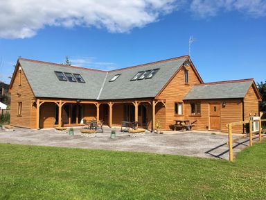 A wooden house with a grey roof and outdoor seating on a paved area with grass in front at Riverside Lodge in Washford