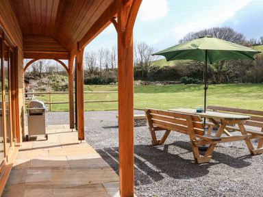 A covered patio with wooden posts a barbecue grill and an outdoor picnic table with a green umbrella in a grassy yard at Riverside Lodge in Washford