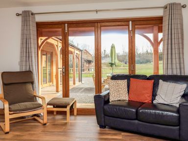 A living room with a brown armchair and footstool next to a black leather sofa with cushions and wooden framed glass doors leading outside at Riverside Lodge in Washford