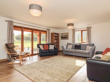 A living room with sofas armchair bookshelf rug and large windows overlooking a garden at Riverside Lodge in Washford