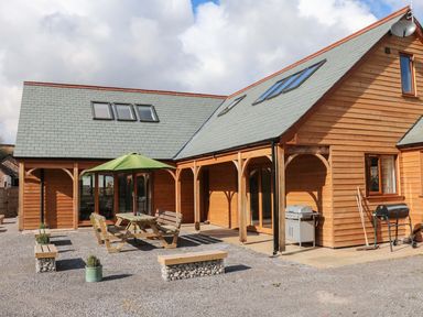 An outdoor seating area with wooden benches and table under a green umbrella next to a wooden house with a gray roof and barbecue grills at Riverside Lodge in Washford