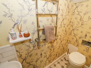 A bathroom with a toilet a towel rail a small shelf with toiletries and floral bird patterned wallpaper at Lower Pack Horse Cottage in Winchcombe