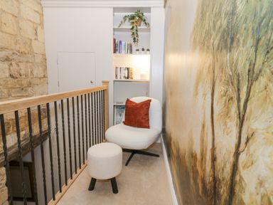A small reading nook with a white chair and footstool near a bookshelf and a mural on the wall at Lower Pack Horse Cottage in Winchcombe
