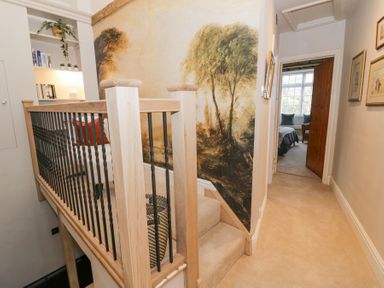 A hallway with a carpeted floor a staircase with wooden and metal railing a wall mural of trees and a view into a bedroom at Lower Pack Horse Cottage in Winchcombe