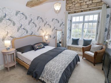 A bedroom with a bed bedside tables lamps armchair window with cushions and peacock wallpaper at Lower Pack Horse Cottage in Winchcombe