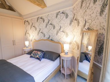 A bedroom with a bed a decorative pillow two bedside tables lamps a full length mirror and peacock patterned wallpaper at Lower Pack Horse Cottage in Winchcombe