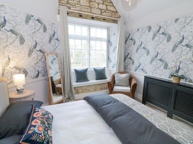 A bedroom with a bed a window seat with pillows a wicker chair and a full length mirror at Lower Pack Horse Cottage in Winchcombe