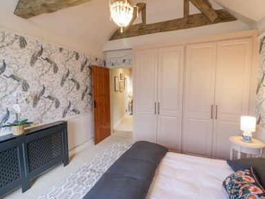 A bedroom with a bed a bedside table with a lamp a wardrobe and floral wallpaper at Lower Pack Horse Cottage in Winchcombe