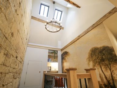 A hallway with stone and painted walls a hanging chandelier and a built in bookshelf with books at Lower Pack Horse Cottage in Winchcombe