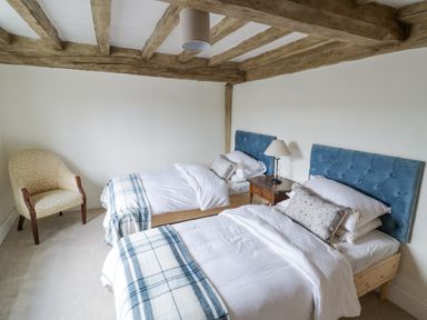 A bedroom with two single beds with blue headboards a wooden bedside table with a lamp and a beige armchair at Lower Pack Horse Cottage in Winchcombe