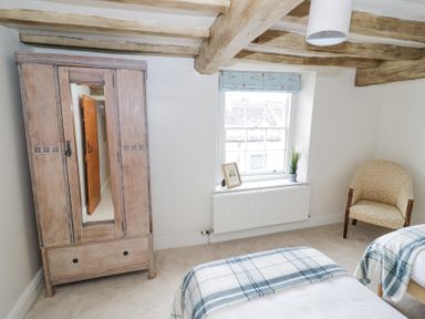 A bedroom with wooden wardrobe window with plants and picture two beds with plaid blankets and an armchair at Lower Pack Horse Cottage in Winchcombe