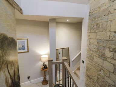 A stairway landing with a wooden banister a small round table with a lamp and a framed picture on the wall at Lower Pack Horse Cottage in Winchcombe