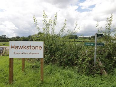 A sign for Hawkstone brewery shop taproom next to public footpath signs in a grassy rural area