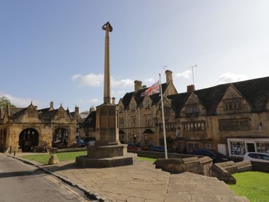 A stone monument and union jack flagpole in front of historic buildings in a town square at Lower Pack Horse Cottage in Winchcombe