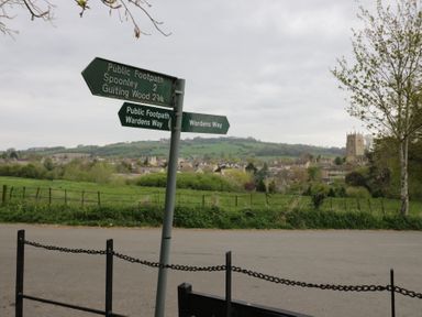 A road with signposts for public footpaths and a village with a church and hills in the background at Lower Pack Horse Cottage in Winchcombe