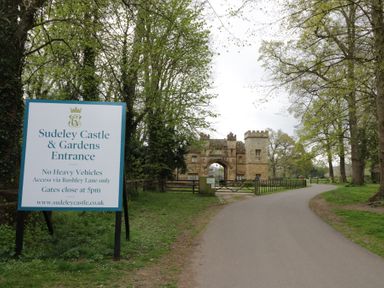 A pathway leading to a stone archway with a gate and a sign for Sudeley Castle and Gardens Entrance surrounded by trees