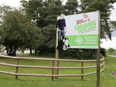 A wooden fence and a sign for Adam Henson's Cotswold Farm Park with trees in the background