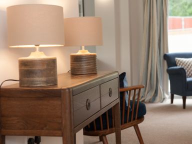 A wooden side table with two table lamps next to a wooden chair and a blue armchair in a room at Brooklands in Uplyme