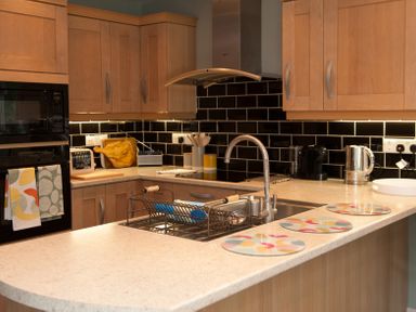 A kitchen with wooden cabinets a black tiled backsplash a sink with a drying rack and patterned placemats on the counter at Brooklands in Uplyme