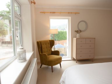 A bedroom corner with a mustard armchair next to a window and a wooden chest of drawers with a round mirror above at Brooklands in Uplyme