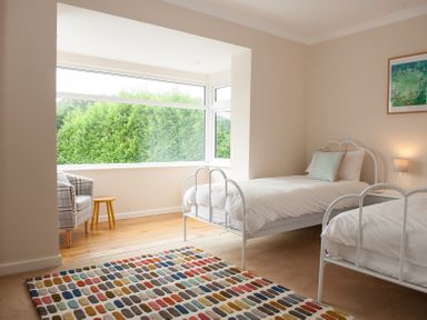 A bedroom with two single beds a colorful rug a plaid armchair and a small wooden stool by a large window at Brooklands in Uplyme