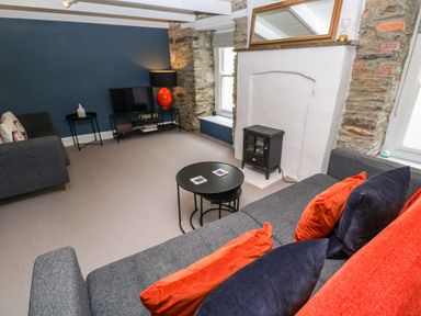 A living room with grey sofas orange and dark blue cushions a black round coffee table a small black stove a television on a stand and a stone wall at Winterbrook Cottage in Mevagissey