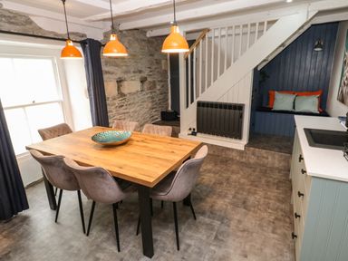 A dining area with a wooden table and six chairs under orange pendant lights and a seating nook under stairs at Winterbrook Cottage in Mevagissey