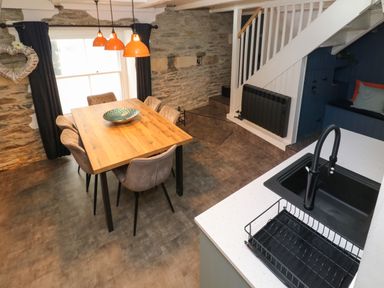 A dining area with a wooden table and six chairs under orange pendant lights next to a kitchen sink and stairs with stone walls at Winterbrook Cottage in Mevagissey