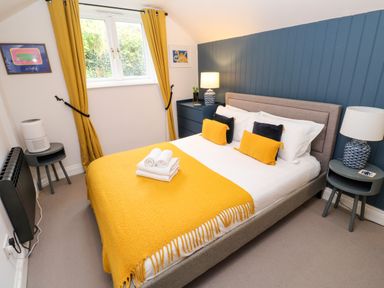 A bedroom with a bed with yellow blanket and cushions beside blue wall and window with yellow curtains at Winterbrook Cottage Mevagissey