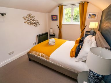 A bedroom with a bed covered in white linens and a mustard yellow throw with matching curtains and wall art at Winterbrook Cottage in Mevagissey