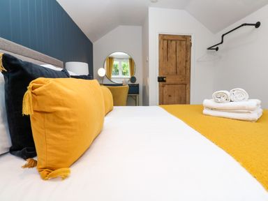 A bedroom with a bed with yellow and dark pillows a wooden door a round mirror on a desk and folded towels on the bed at Winterbrook Cottage in Mevagissey