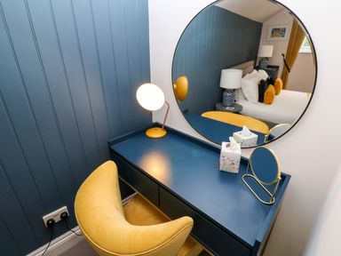 A blue desk with a yellow lamp a yellow chair and a round mirror reflecting a bed at Winterbrook Cottage in Mevagissey