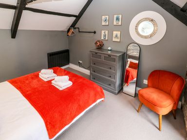 A bedroom with a bed covered by an orange blanket and towels on top a gray chest of drawers a tall mirror an orange chair and framed pictures on a gray wall at Winterbrook Cottage in Mevagissey