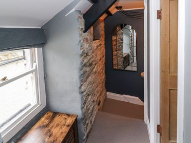A narrow hallway with exposed stone wall a wooden cabinet a window and a large mirror at Winterbrook Cottage in Mevagissey