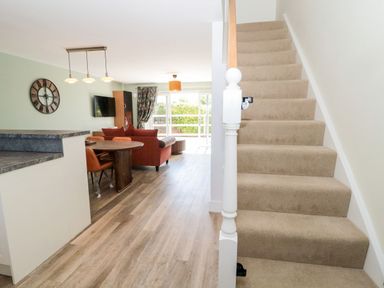 A living room with a dining table and chairs a wall clock a television a sofa and carpeted stairs at Rose Lodge in High Newton-by-the-Sea near Embleton
