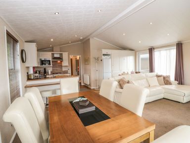 An open-plan living space with a wooden dining table and white chairs a kitchen area with white cabinets and appliances and a white sectional sofa near windows at Walkers Rest in Warton near Carnforth