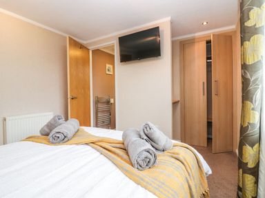 A bedroom with a bed covered with a yellow striped blanket and rolled gray towels a wall mounted tv an open wooden door and an open wooden wardrobe at Walkers Rest in Warton near Carnforth