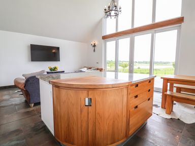 A kitchen island with wooden cabinets and granite countertop in a room with large windows a wooden dining table and sofas at Polly's Annexe in Levington