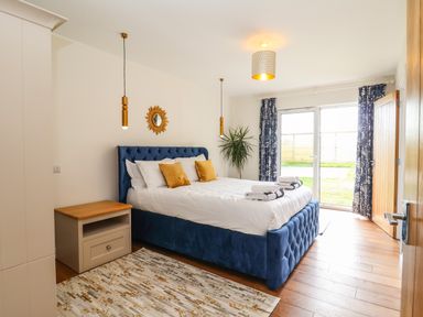 A bedroom with a blue upholstered bed with white bedding and yellow pillows a nightstand rug plant and glass doors with curtains at Pollys Annexe in Levington