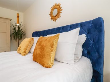 A bed with white sheets two gold pillows a blue tufted headboard a pendant light plant and a sunburst mirror on the wall at Pollys Annexe in Levington