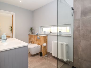 A bathroom with a wall-mounted toilet wooden paneling a white radiator a gray vanity with a sink and a glass shower enclosure at Pollys Annexe in Levington