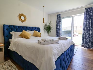 A bedroom with a blue upholstered bed with white bedding and yellow pillows near glass doors with blue curtains at Polly's Annexe in Levington