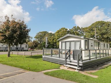 A white static caravan with a gray deck and chairs on a grassy area near a road at The Olive Lodge 227 Sycamore in New Milton