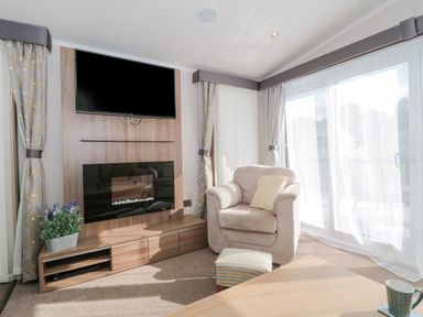A living room with an armchair a footstool a television mounted above a fireplace and a large window with curtains at The Olive Lodge 227 Sycamore in New Milton