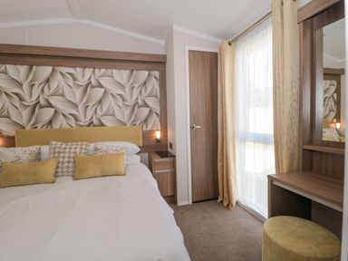 A bedroom with a bed pillows a bedside table a door a window with curtains and a dressing table with stool at The Olive Lodge 227 Sycamore in New Milton