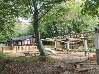 An outdoor playground with wooden picnic tables trees and a fenced wooden building at The Olive Lodge 227 Sycamore in New Milton