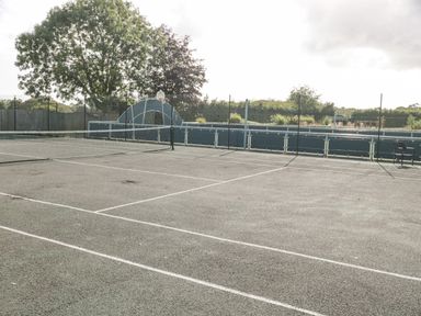 An outdoor tennis court with a net and chain link fence surrounded by trees at The Olive Lodge 227 Sycamore in New Milton