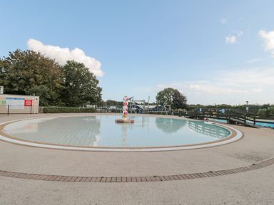 An outdoor shallow pool with a central fountain and playground equipment in the background at The Olive Lodge 227 Sycamore in New Milton