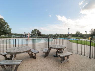 An outdoor area with two wooden picnic tables near a fenced swimming pool and a playground in the background at The Olive Lodge 227 Sycamore in New Milton